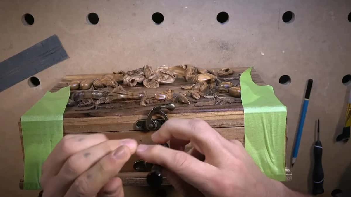 Hands fasten a brass clasp onto a small carved wood box, its sides taped for protection, a ruler and pencil nearby.