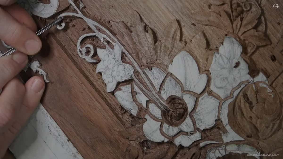 A hand carves fine detail into a wood flower and vine relief, white chalk lines marking the petal edges.