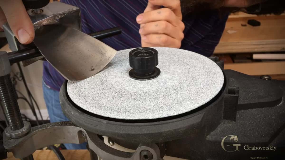 A big gouge held bevel down on a grey abrasive disc, with the guide arm clamped at the left.