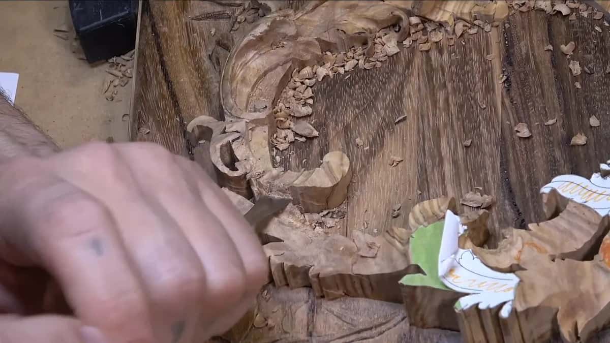 A fist grips a carving tool, mid stroke into a wood scroll relief, with fresh shavings piled around the groove.