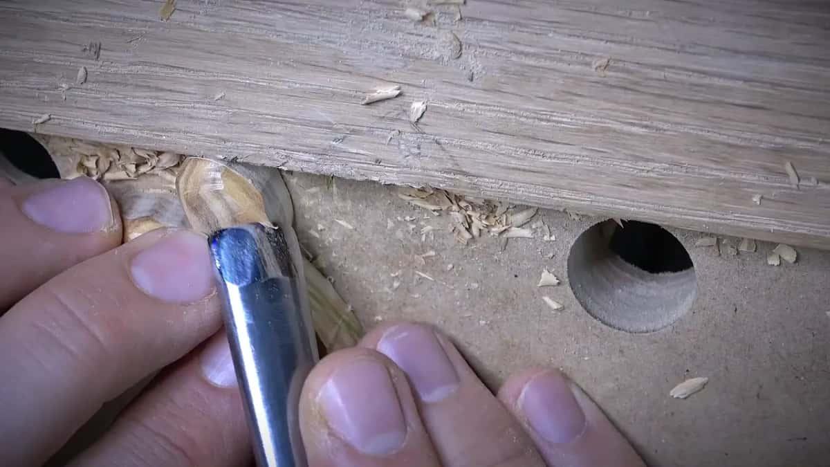Fingers steady a small striped wood shaving while a gouge trims it beside a workbench hole.