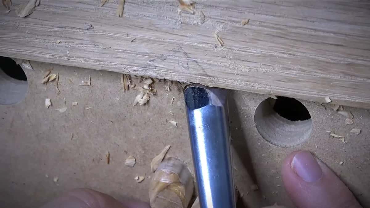 A rounded gouge is held beneath a pale wood board marked with pencil lines and scattered shavings.