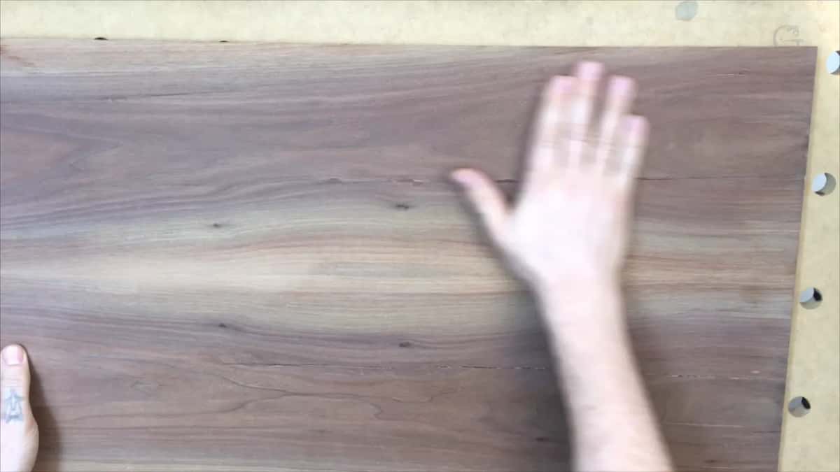 An open hand rests on a wide dark brown board with straight grain and a few small knots.