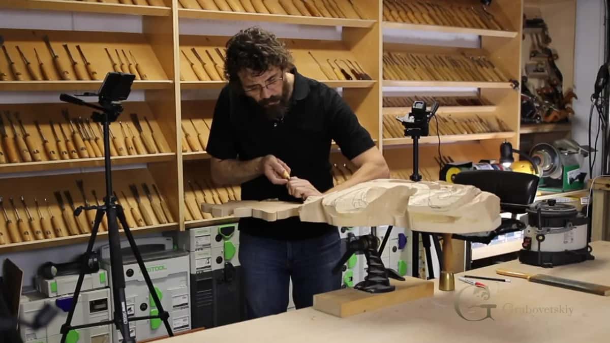 Standing at the bench working a large basswood blank with a small gouge, racks of gouges on the wall behind