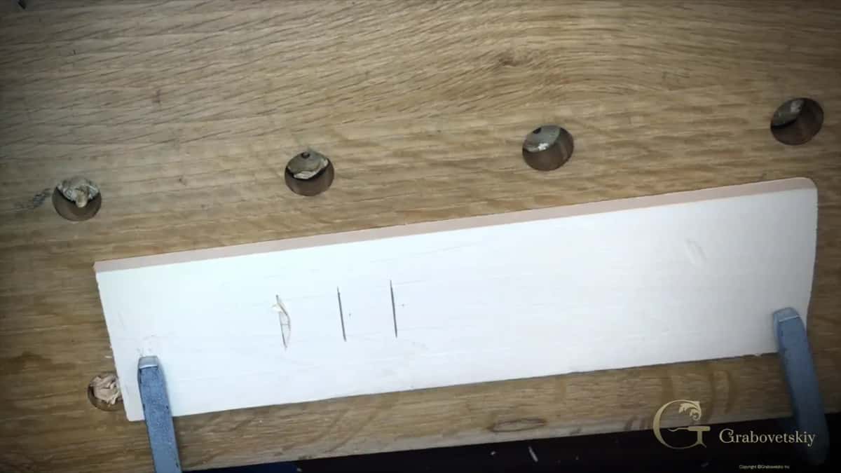 Three straight-down knife slices and one lifted chip on a pale basswood practice board clamped to an oak bench