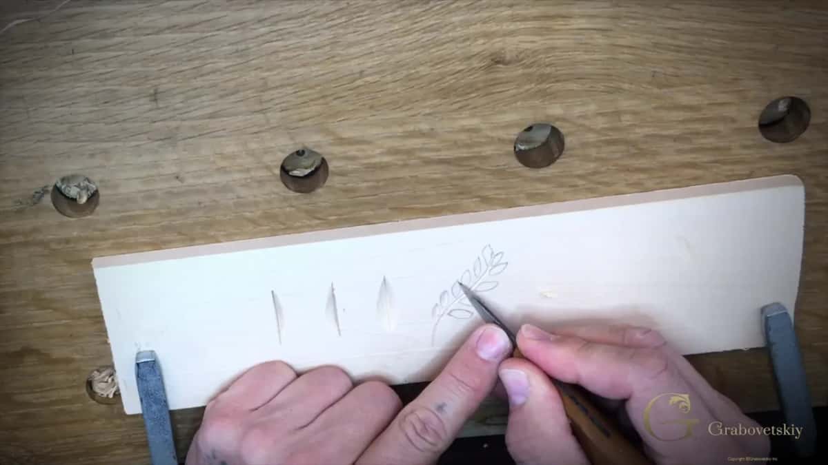 Both hands on a chip carving knife, thumb up near the blade, working a penciled leaf spray on a clamped board