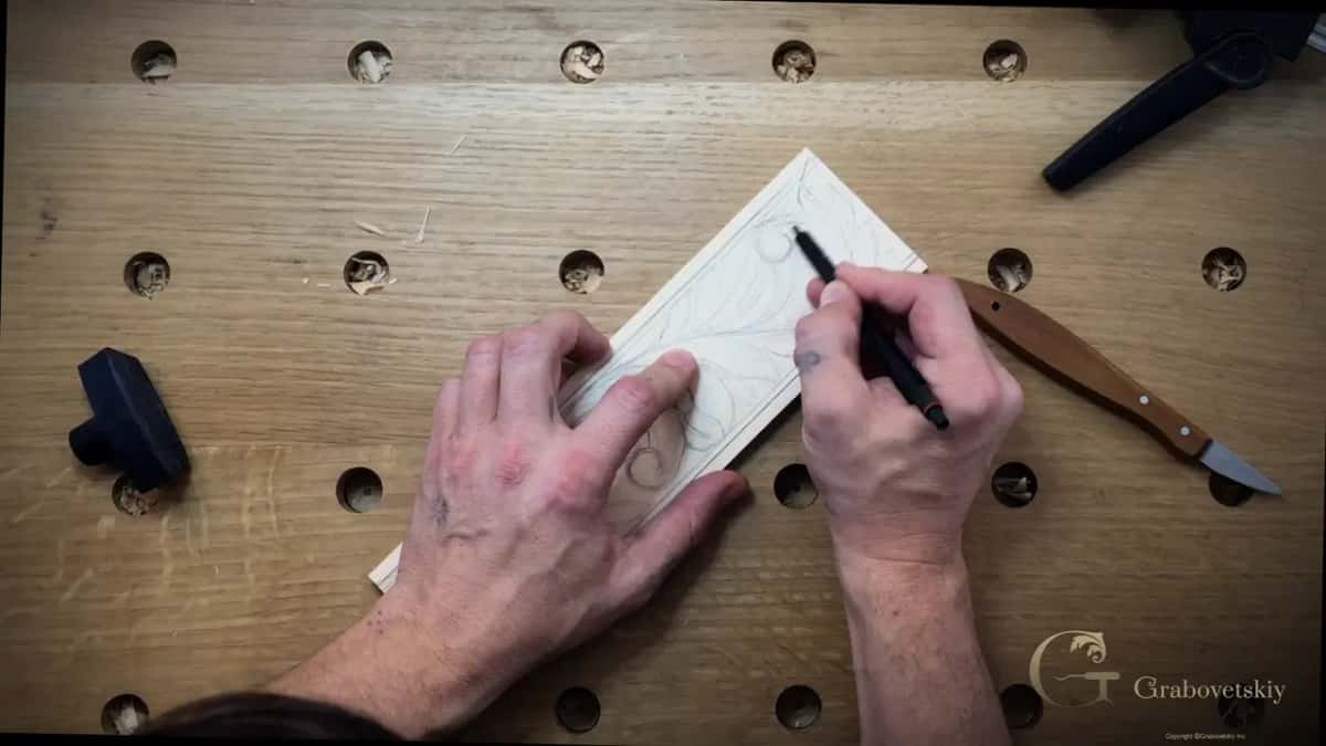 Both hands drawing a pencil scroll on a small basswood board, a chip carving knife and rubber mallet on the bench beside it