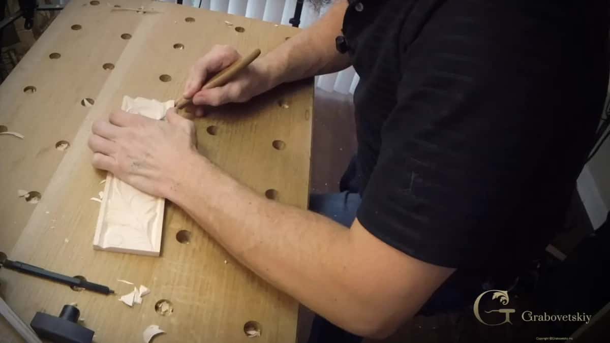 Both hands at the knife tip, left hand planted on a narrow basswood panel with a carved leaf, shavings on the bench