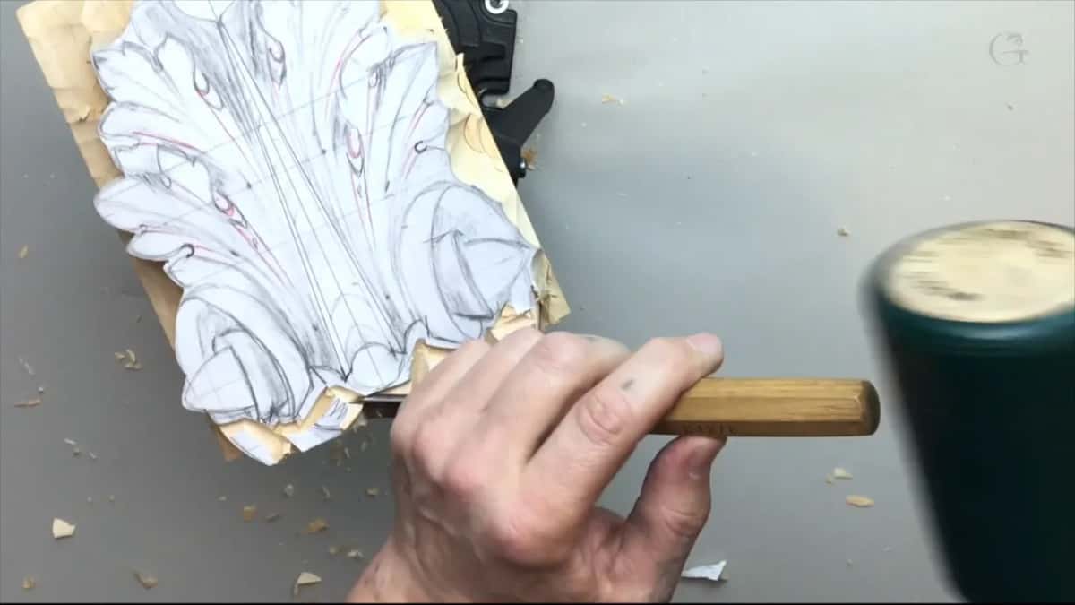 A hand on a stamped gouge handle at the edge of a paper acanthus pattern, a green rubber mallet swinging in from the right