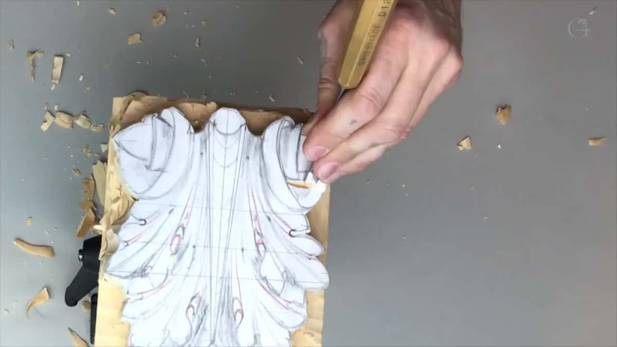 Waste carved away around a paper acanthus pattern, a Swiss made gouge held at the outline, shavings on the grey bench