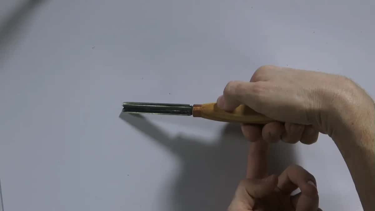 A gouge held blade out against a plain wall, the bevel in profile, the other hand's knuckle just under the handle