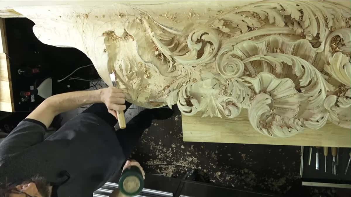 A gouge held against a deeply carved acanthus frieze while the other hand raises a green rubber mallet