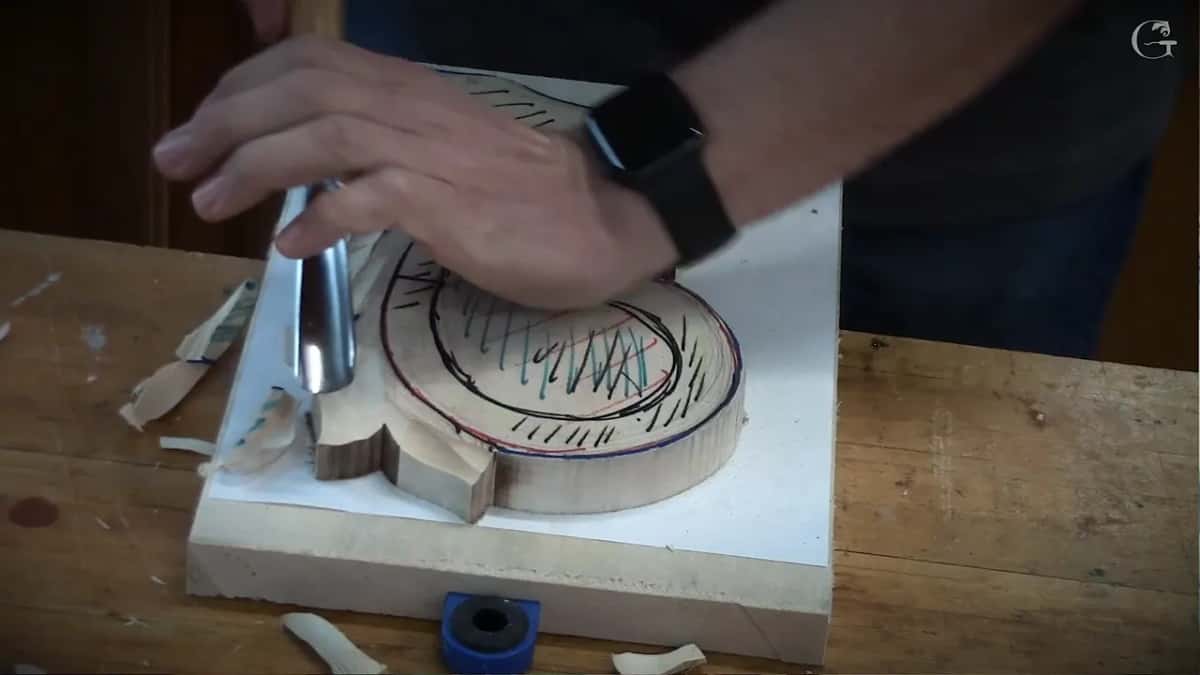 One hand pressed flat on a round basswood blank marked in black, red and teal while a wide gouge sits against its edge
