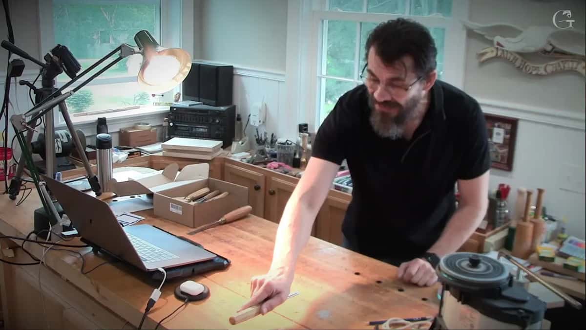 A carving bench in a bright room with a laptop, a lamp, a box of gouges and turned mallets standing at the far end