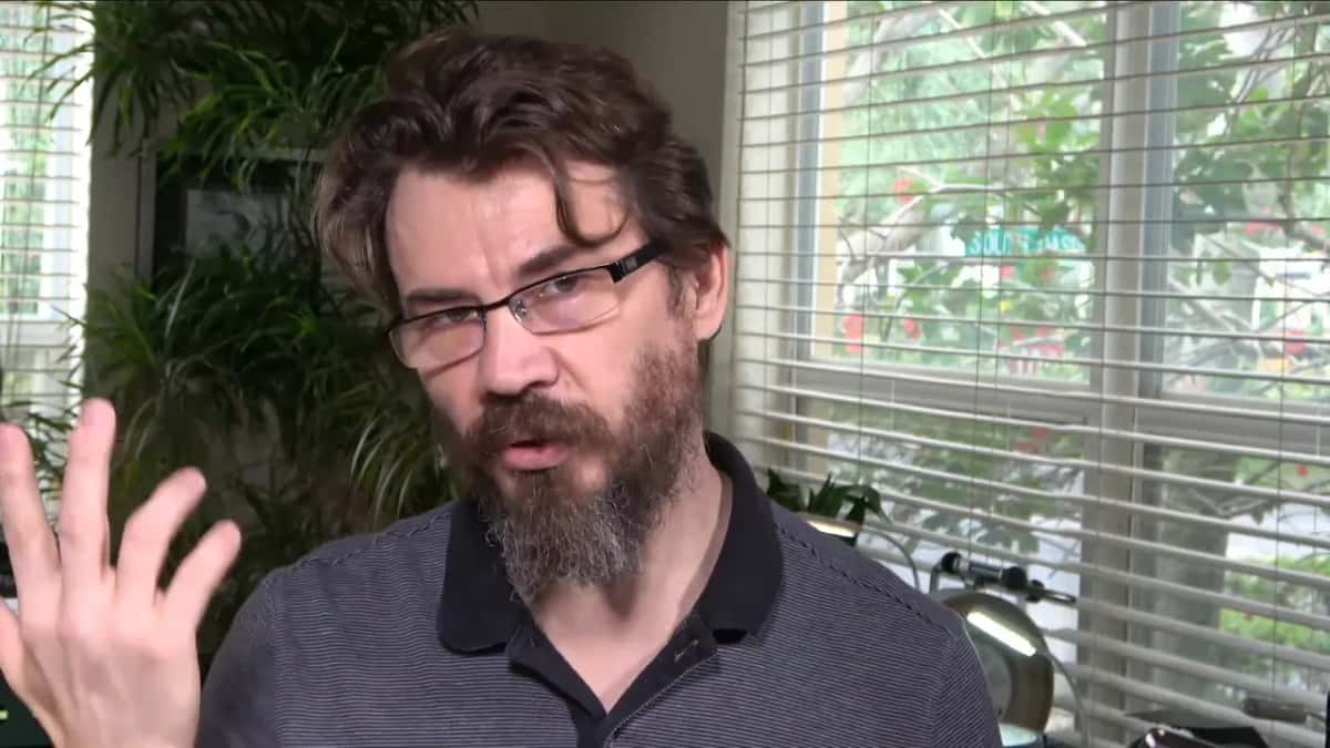 Seated near a window, a bearded man in glasses talks to the camera with one hand raised.