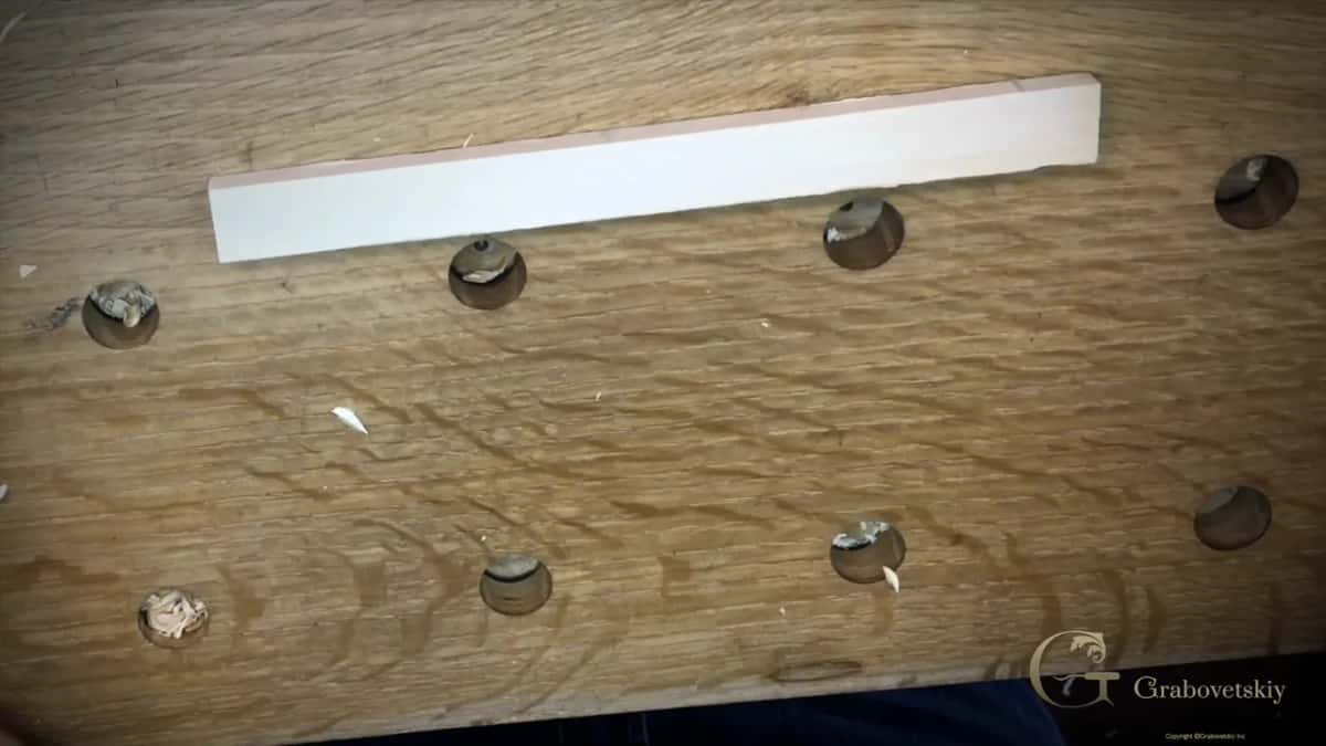 One thin narrow strip of pale wood lying on an oak bench top dotted with round bench dog holes.