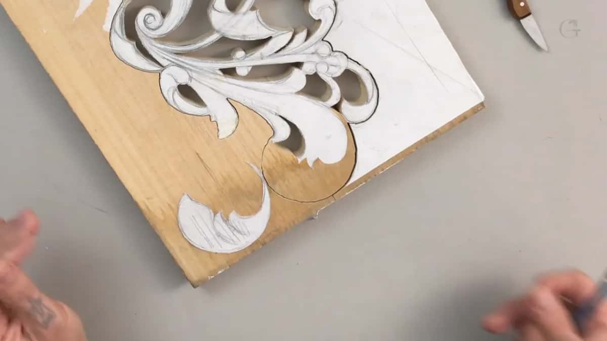 Corner of a small pale offcut with a paper scroll pattern glued down, part of it carved away, a knife lying beside it.