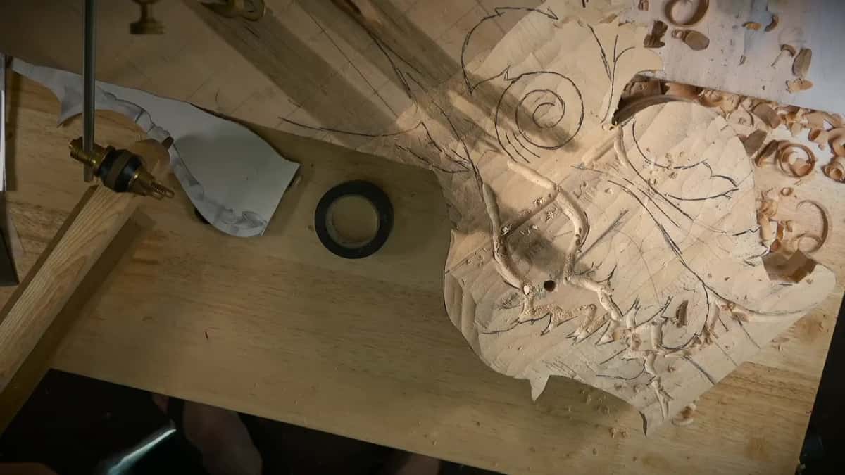 A small sawn out scroll blank lies on a bench board beside a paper pattern, a tape roll and the post of a hold down.