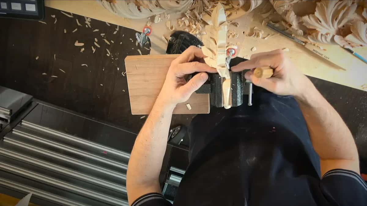 Seen from above, a small carved leaf gripped in a bench vise while both hands adjust it, shavings scattered behind.