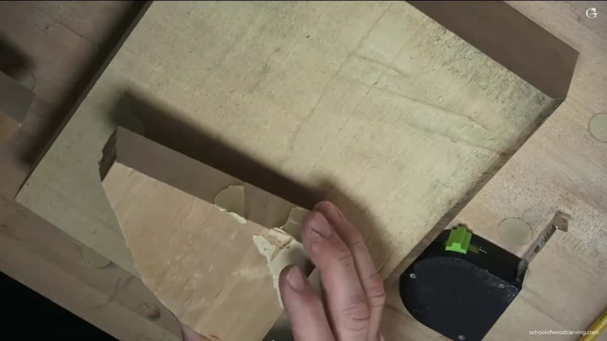 A hand holds a small pale offcut against a larger board on the bench, a clamp head just visible at the right.