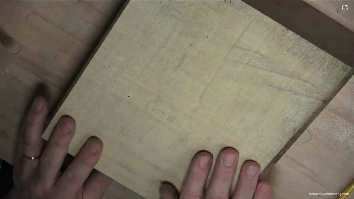 A small square blank of pale wood held flat between two hands on the bench, its grain and saw marks showing.