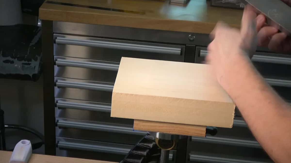 A small pale offcut block mounted on a carving post at bench height, a hand moving above it with a tool.