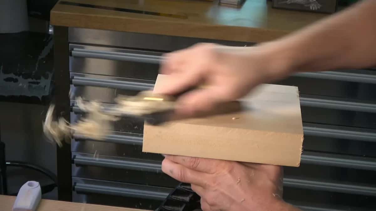 Shavings spray off the top of the same small offcut block as a hand drives a tool across its face.