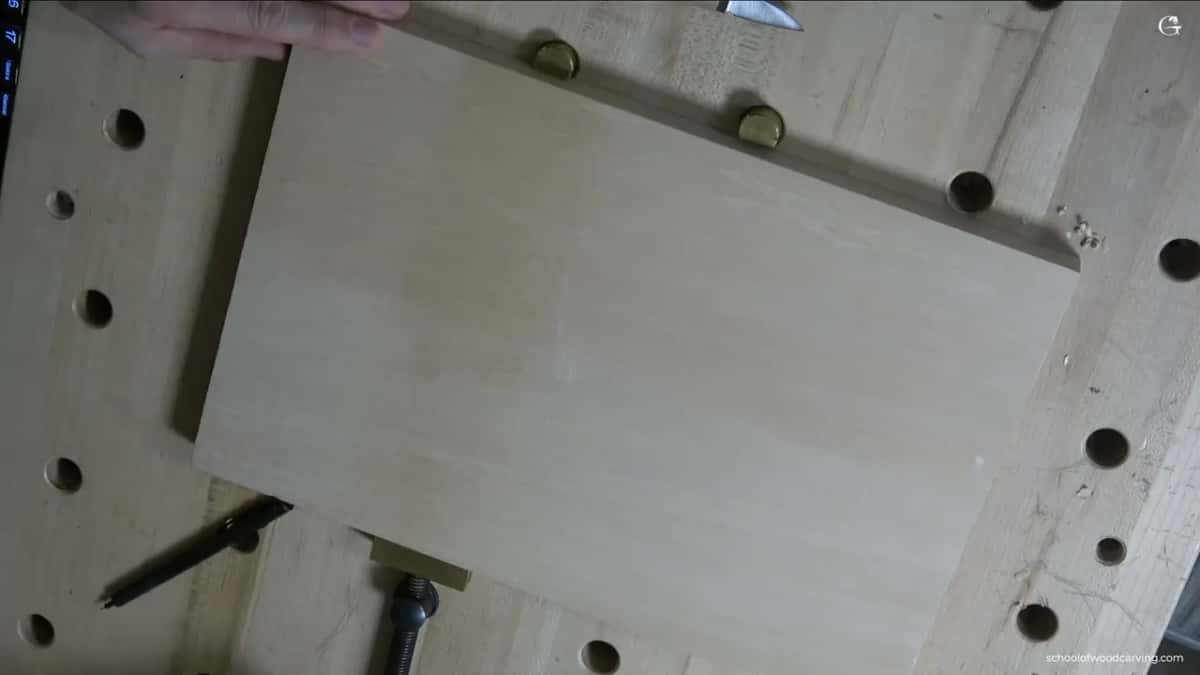 A clean pale board held down between brass bench dogs, a pencil and a knife waiting beside it before any carving starts.
