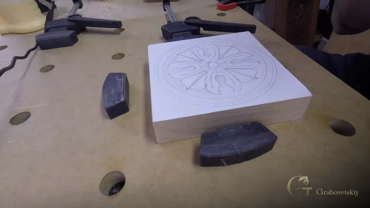 A small square block primed white with a rosette drawn on it, resting on the bench between two dark clamp pads.