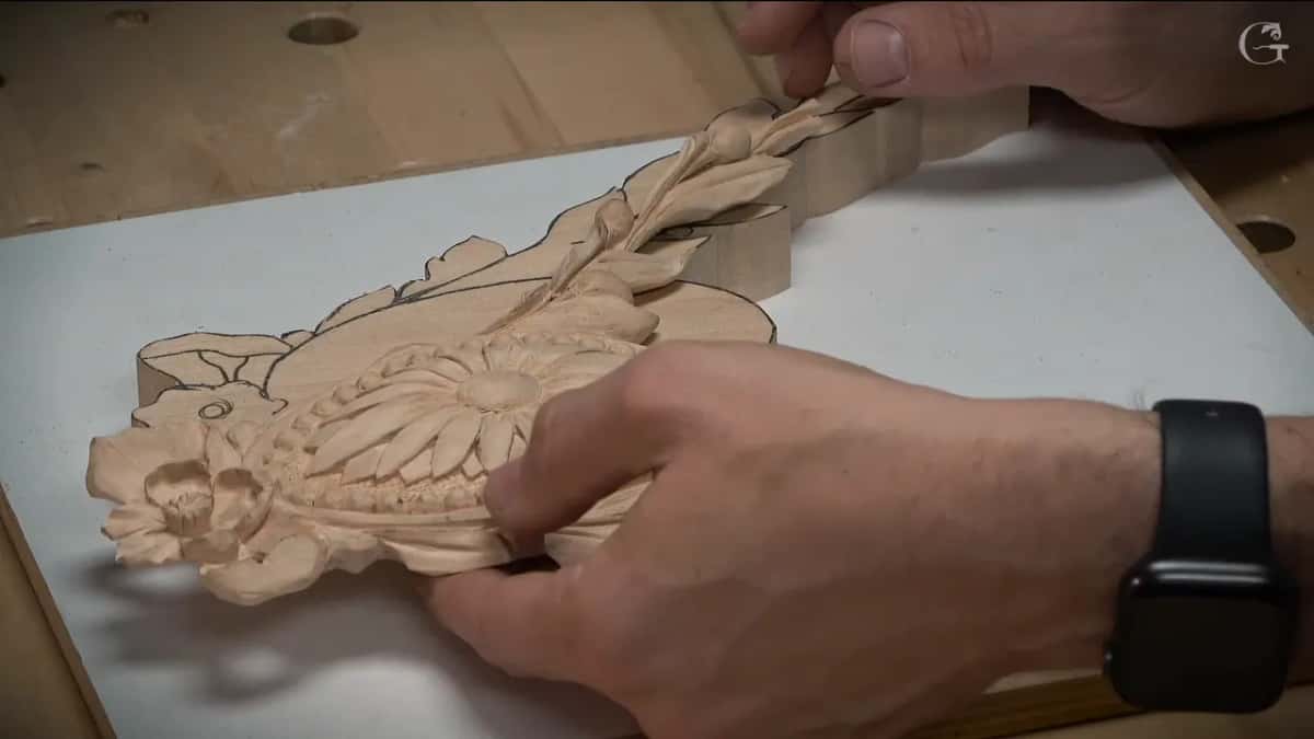 Hands tilt a small sawn out piece so the carved daisy and its ribbon of leaves catch the light, its thin edge showing.