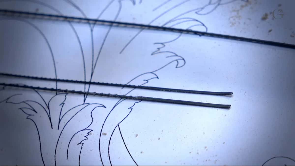 Two fine scroll saw blades lying across a pencil drawing of acanthus leaves on white paper.