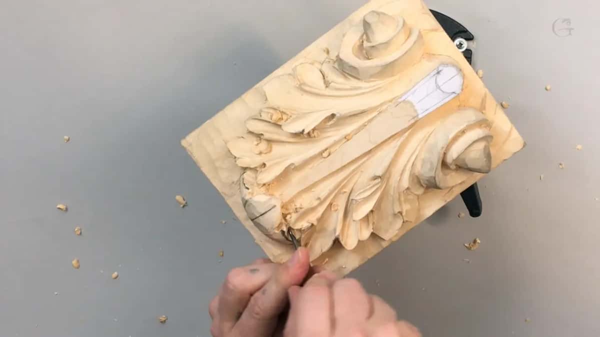 A small board clamped at one edge carries a full acanthus leaf, hands bringing a fine tool up to it from below.