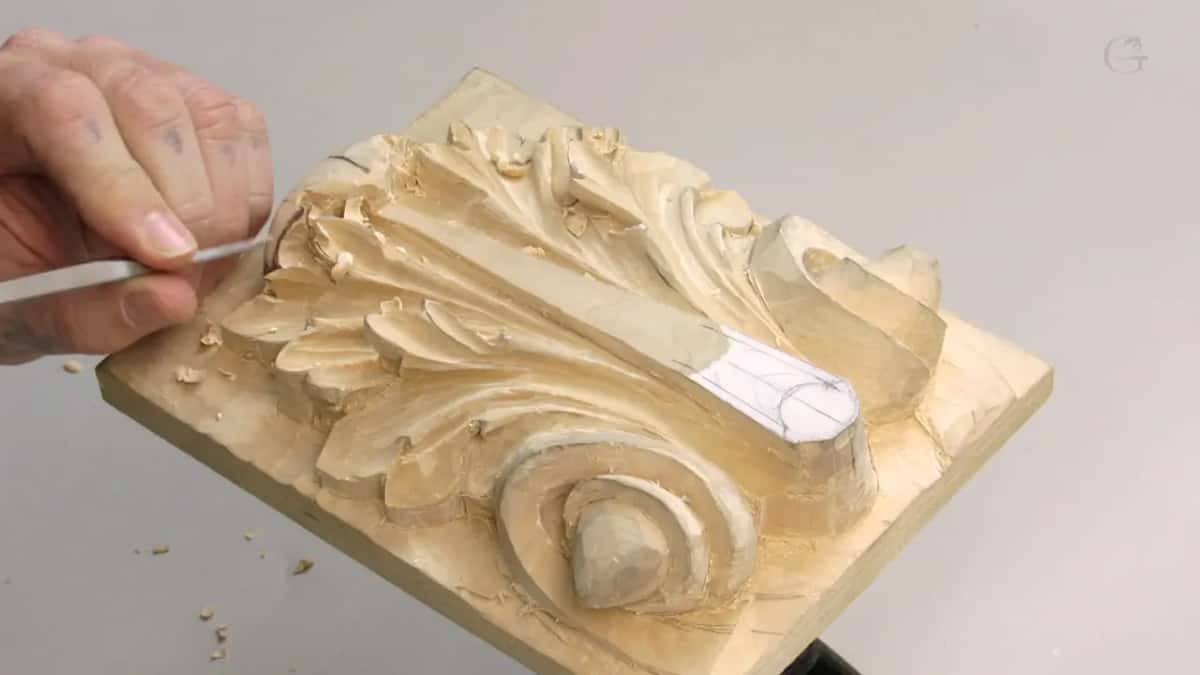 Closer and from the side, one hand steadies a slim tool against the deep leaf carved into that small clamped board.