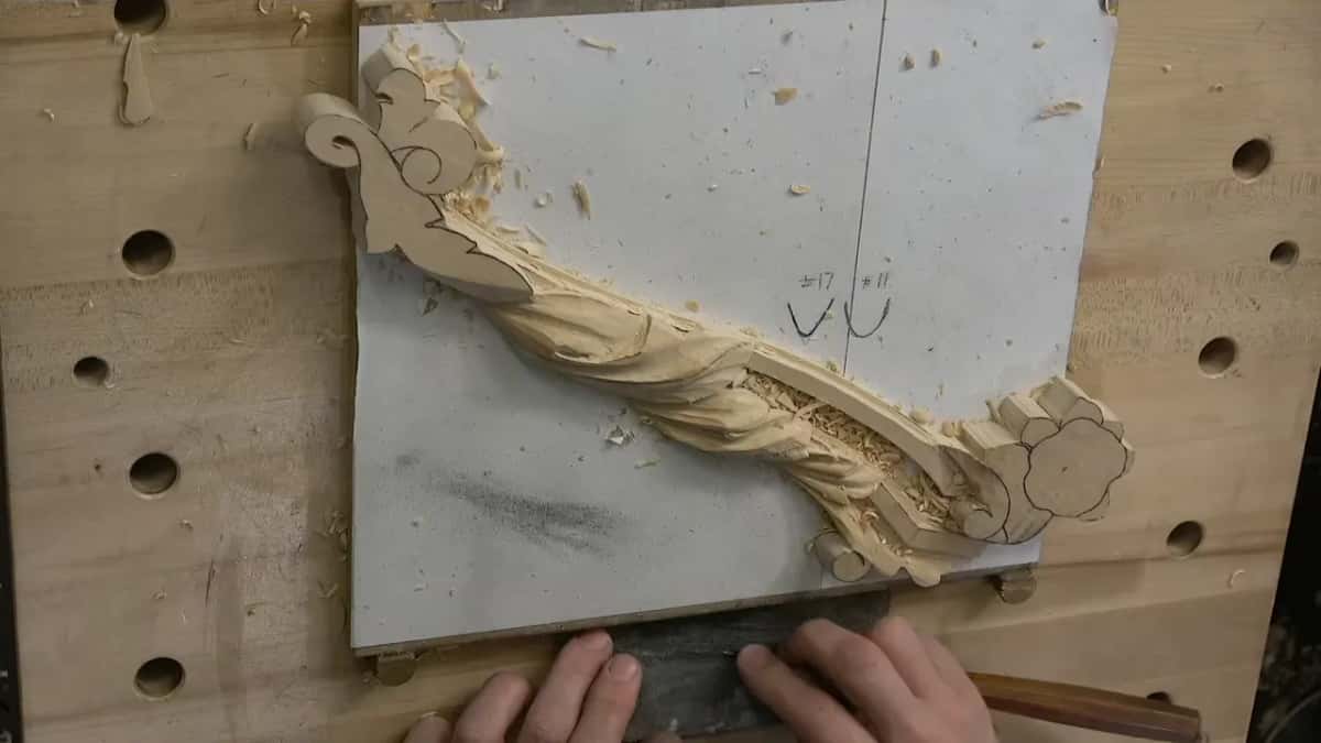 Overhead view of a thin pale board pinned between bench dogs, a carved scroll running across it under loose chips.