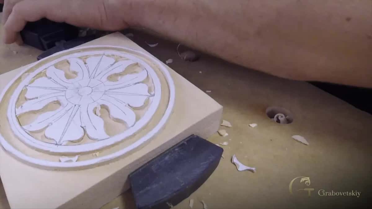 A small rosette part carved into its own little square blank, chips lying around it and a dark caul at one edge.