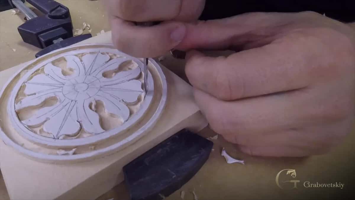 Both hands steer a slim tool into the petals of that same small rosette, a bar clamp holding the blank from the left.