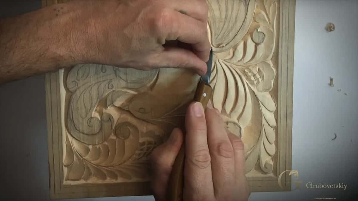 A thin carving knife shapes a symmetrical leaf and scroll design into a flat wood panel.