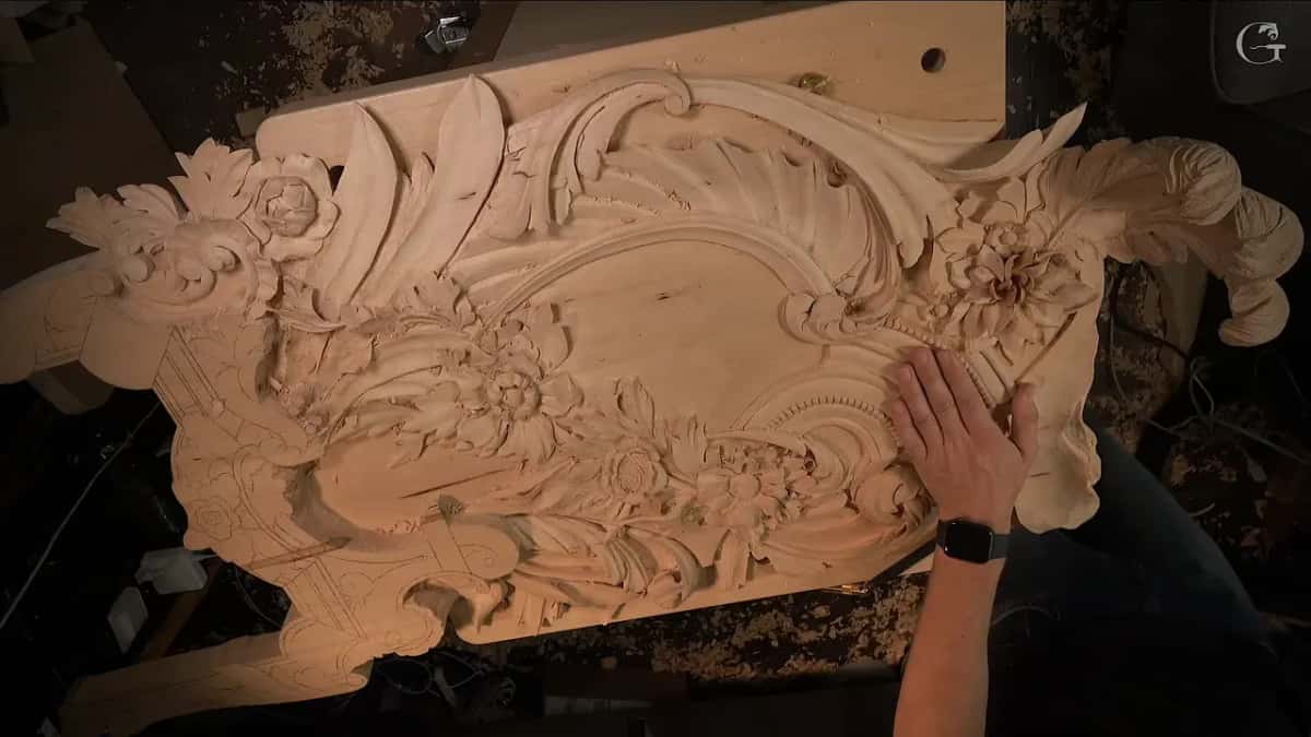 An elaborate wood ornament carved with flowers and scrolling leaves sits on a dark table, a hand resting on its surface.