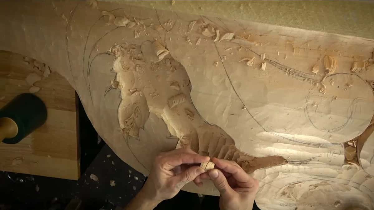 Two hands hold a small wood chip over a large relief carving of roughed-out leaf shapes, shavings covering the surface.