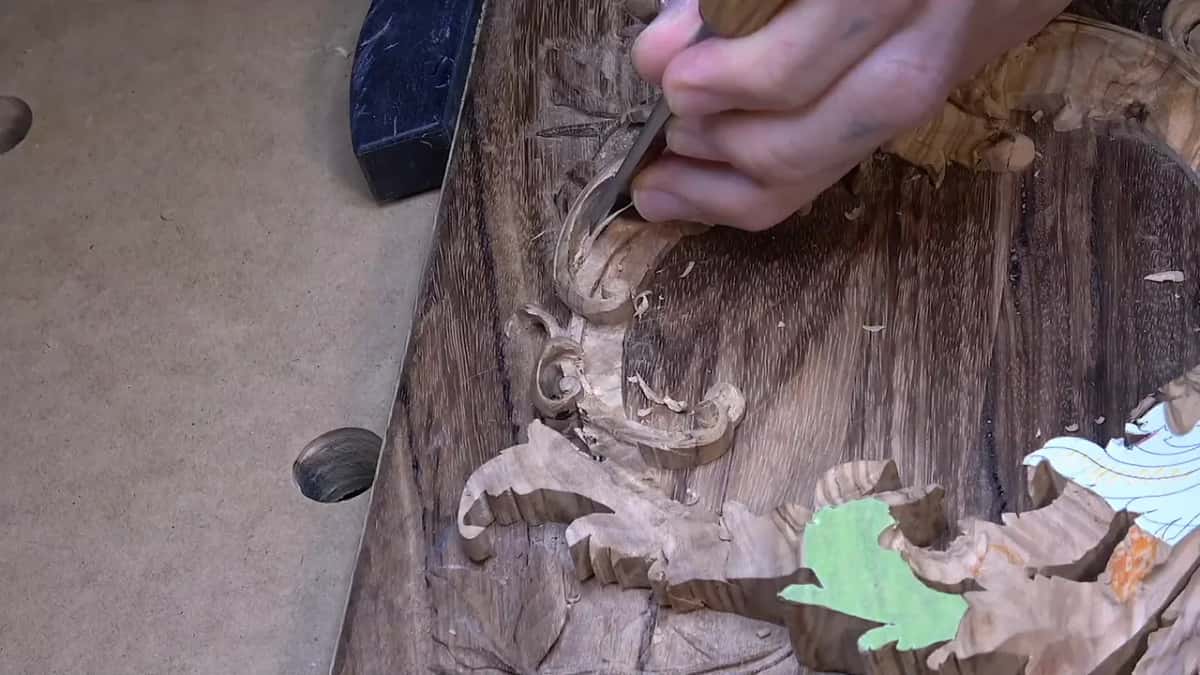 Seen from above, a gouge works leaves into a dark weathered plank, a colored paper pattern beside it.