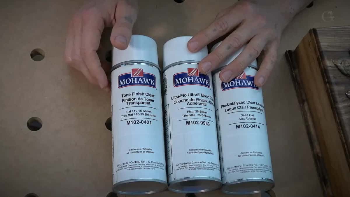 Three Mohawk spray cans of clear finish stand in a row, hands resting on the bench behind them.