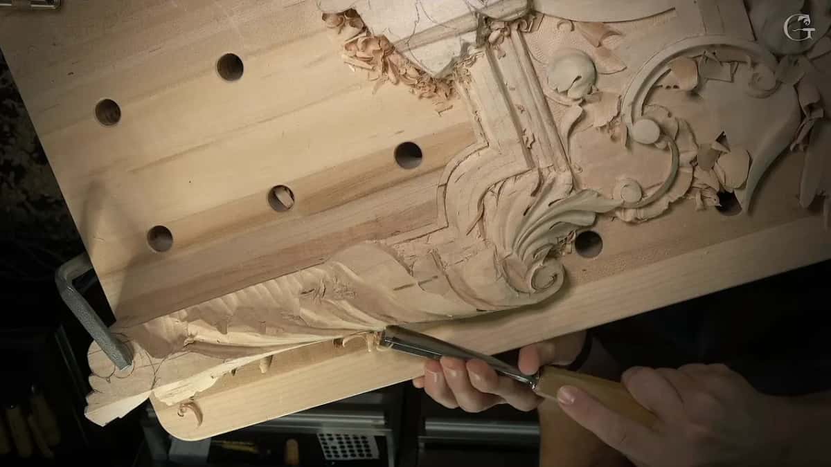 Tilted view of a long panel, part carved and part still flat, a gouge held down at the edge.