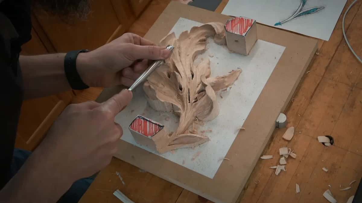 Both hands bring a gouge to that roughed leaf, with a drawing of it lying on the bench nearby.