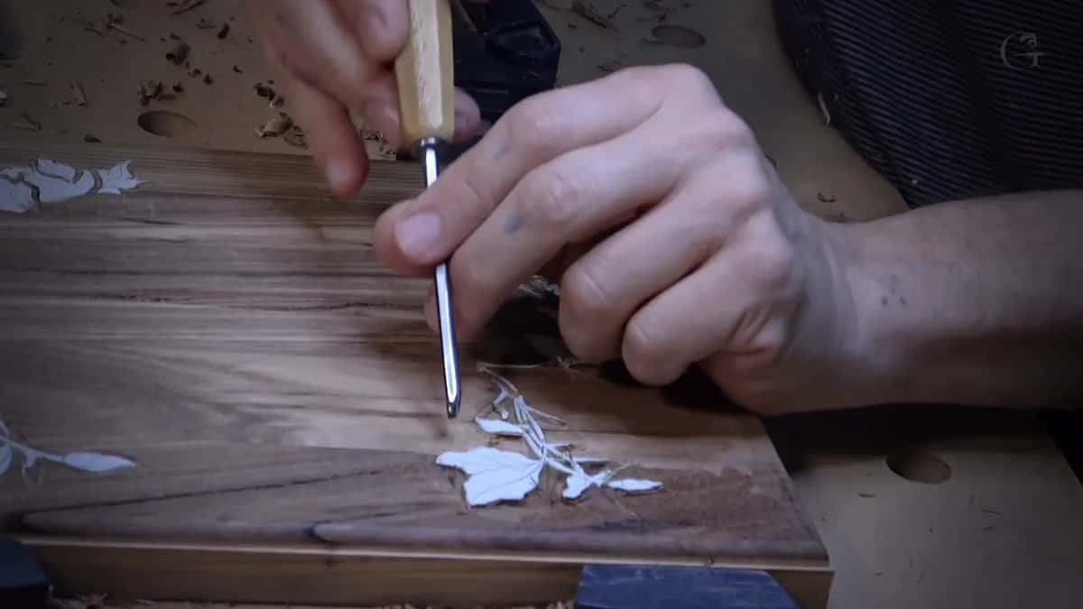 Gouge carving a leaf and vine pattern into a wood board with pencil layout lines still showing.