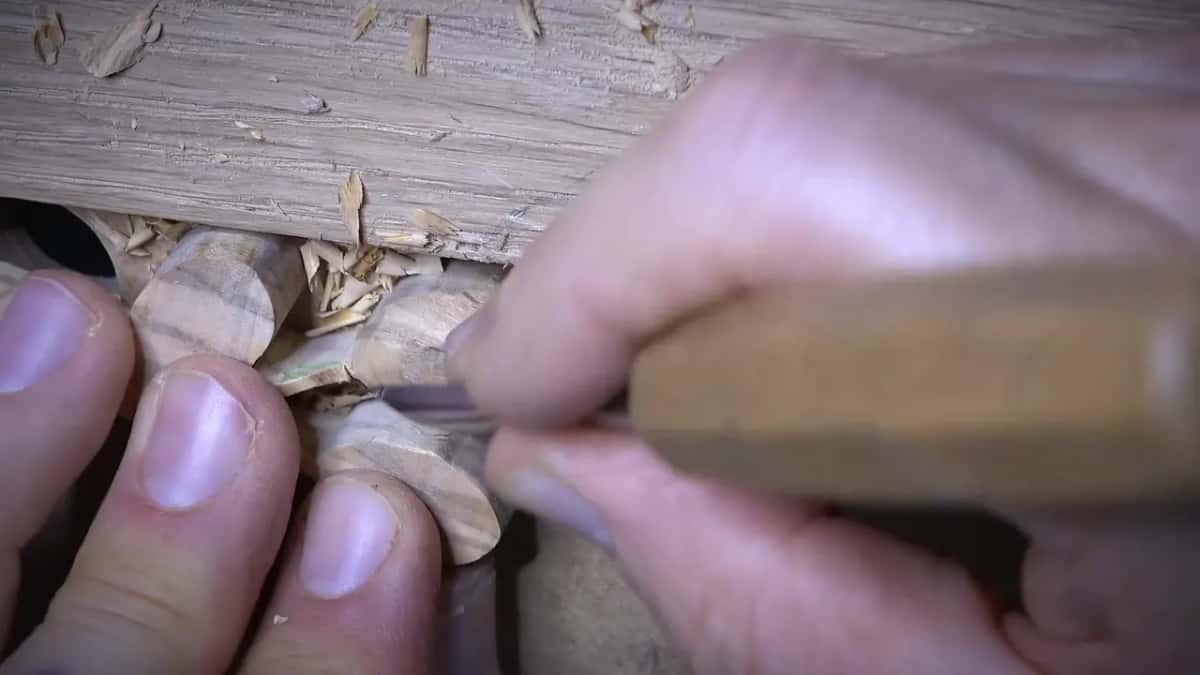 Fingers steady a small pale wood block while a carving tool peels a thin curl of shaving from its edge.