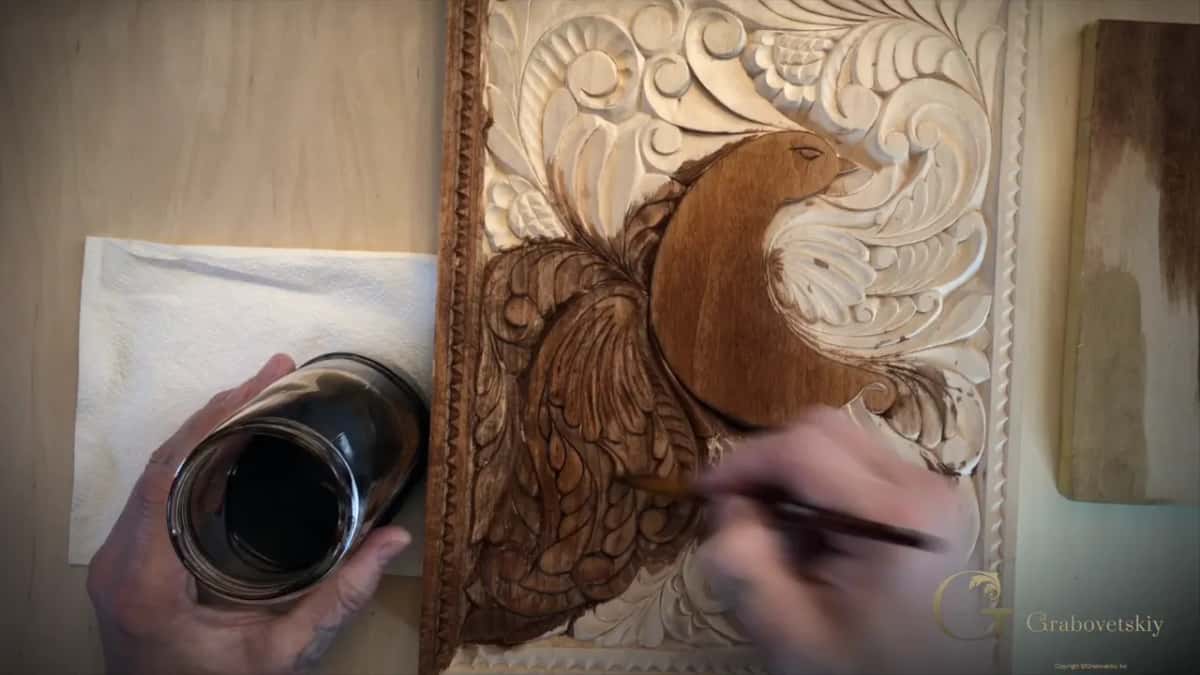 A carved bird panel half stained warm brown and half still pale, with the jar of color held alongside.