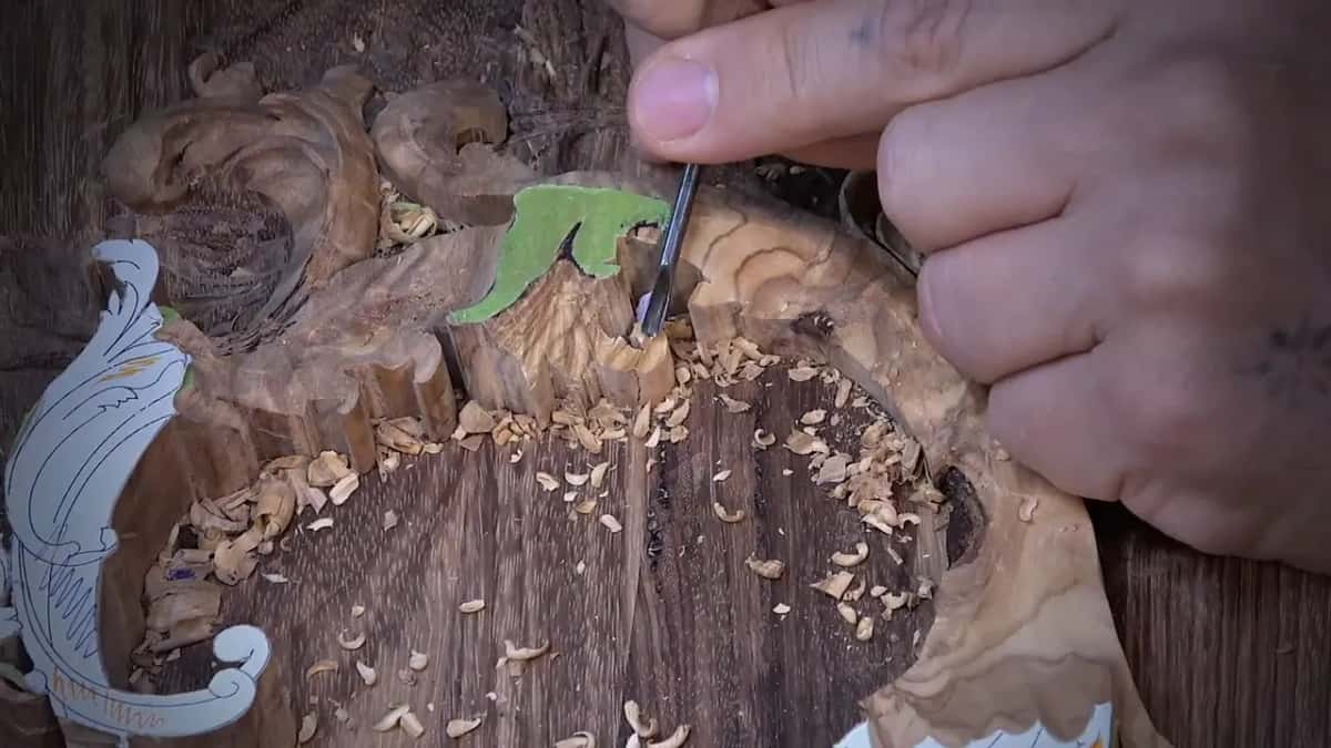 A small tool carves into a dark scroll shape marked with green paint, a paper pattern resting at the edge.