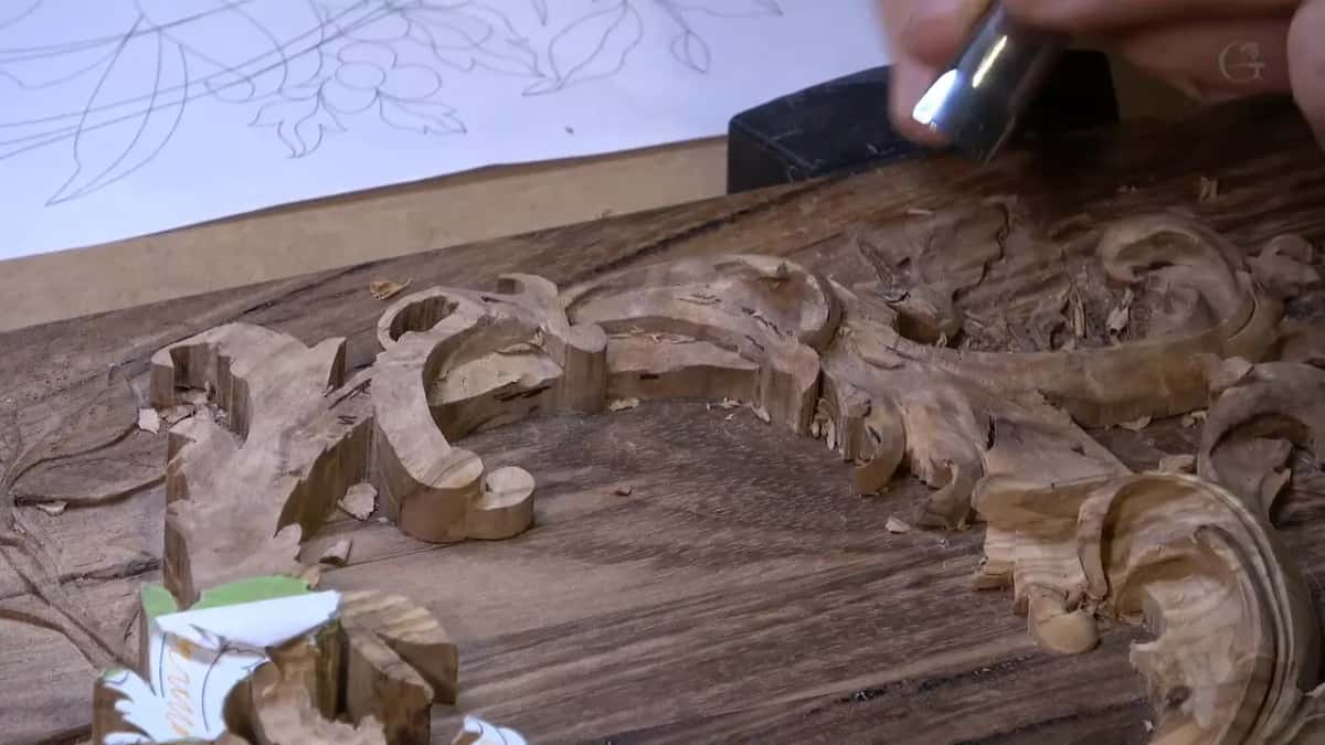 A gouge tip works the edge of a carved scroll pattern, a penciled floral sketch waiting on the paper behind it.