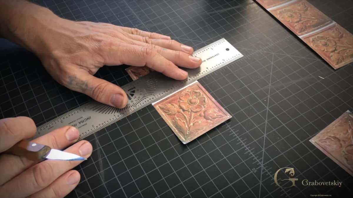 One hand pins the rule while the other brings a slim blade to the edge of a printed flower tile.