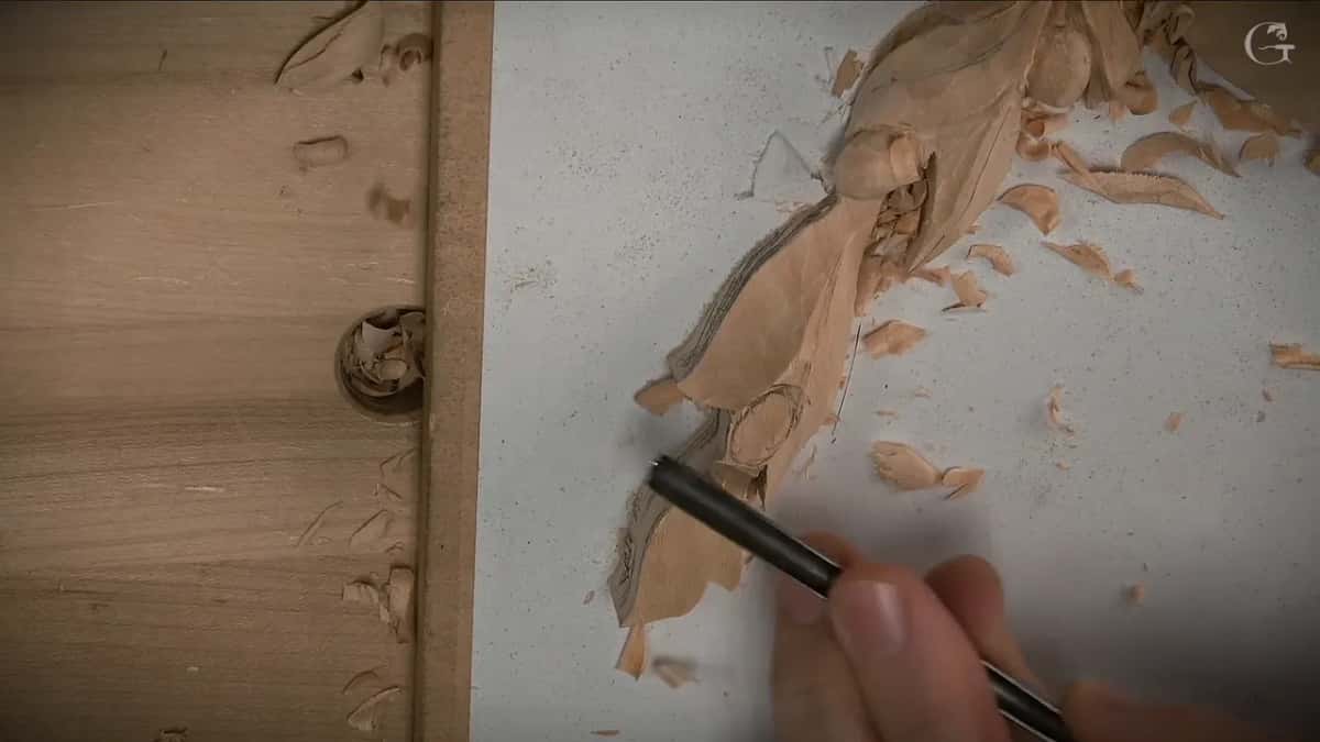 A black pencil comes in low to draw a new line on a carved leaf, loose chips lying all around it.